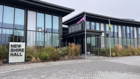 A modern glass-fronted building with the words 'New Shire Hall' on a stone to the right. There are two flag poles, one flying the Union flag and the other the flag of Ukraine. 