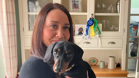 Bella the dachshund in the arms of her owner Debbie , who's got brown hair and is wearing a black jumper 