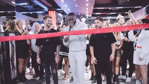 The Couture Club There is a large red ribbon with The Couture Club branding across the front door of The Trafford Centre store with Ross and Scott, and many customers, friends and family, standing behind them. 