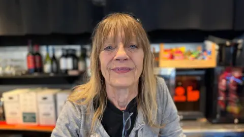 BBC A lady is wearing a black shirt with a grey hoodie. She has blonde hair and is stood in front of a bar.