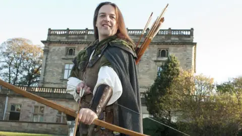 A man with long hair and green robes holding a bow and arrow. He stands in front of Nottingham Castle