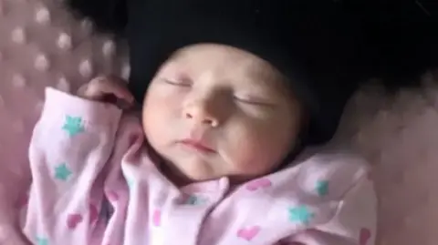 A baby girl wearing pink babygrow and a black woolly hat. 