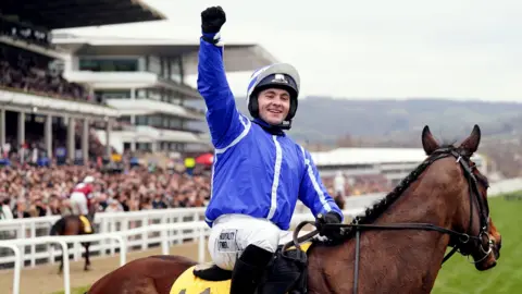 PA Media Jonjo O'Neil wearing a blue jacket, white slacks and a blue helmet, sits on horse Poniros with one arm raised in the air in celebration