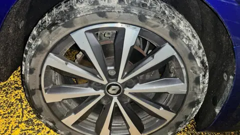 Adrian Hargreaves A close-up view of a damaged wheel attached to a blue vehicle. The alloy wheel has a multi-spoke design in silver and dark grey, with a visible brake disc and caliper behind it. 