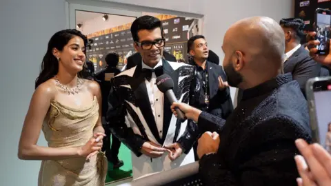 Janhvi Kapoor, dressed in a golden glittery dress, and Karan Johar wearing a white and black tux with a black bow tie and dark rimmed glasses, speaking to Asian Network's Haroon Rashid, who has a dark microphone outstretched. Haroon is wearing a dark suit.