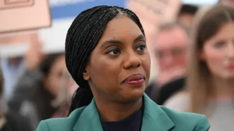 A woman with braided hair wearing a green blazer looks to the side. She appears to be in a crowd, with people and signs blurred in the background.
