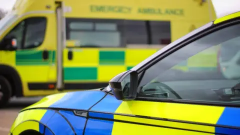 Stock image of the front end of a blue and yellow check police car taken from the passenger side. Part of the interior is visible through the door window. There is a black wing mirror and the picture cuts off just below the top of the wheel arch. A green and yellow check ambulance is parked side-on and appears blurred in the background.