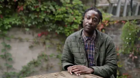 Jeanperry Mundabi is sat outside at a wooden table, in front of a wall covered in plants. He is wearing a green coat and purple checked shirt.