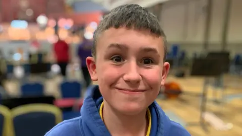 A boy with short brown hair smiling at the camera. He is wearing a blue hoodie with a yellow t-shirt underneath.