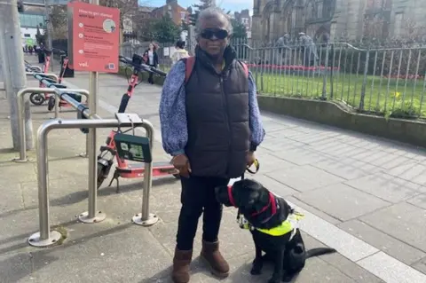 Claire Hamilton/BBC Visually impaired Naomi Ditchfield in black jeans, with a blue patterned blouse and black body warmer with her black labrador guide dog next to an e-scooter stand