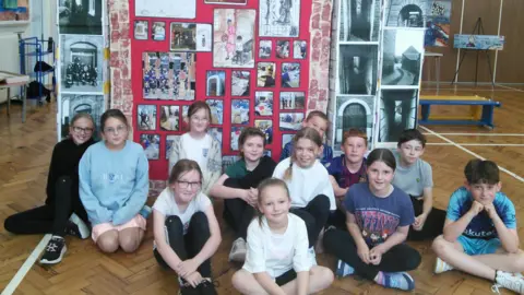 Twelve primary school children sitting on a wooden herringbone floor in front of large display of artwork.