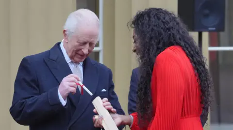 PA Media King Charles wearing a dark suit and grey tie places a message into a wooden baton. A woman with dark curly hair and a red dress holds the baton. 