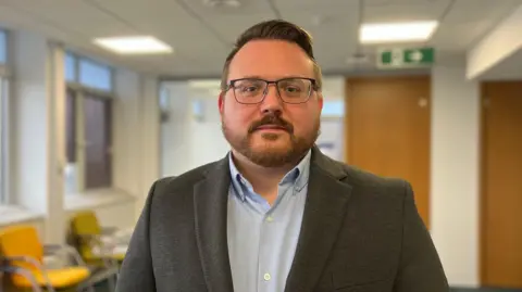 A man in a blue shirt and grey suit jacket. He is standing inside. He is wearing glasses. He has a beard and short hair, but with a quiff. 