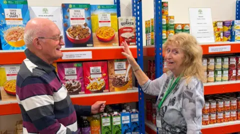 Supplied A man and a woman stand in front of shelves stocked with food items, appearing to be in a food bank or pantry. They are both smiling, and the woman is gesturing towards the shelves. 