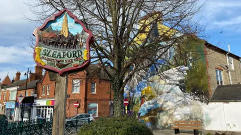 North Kesteven District Council A wooden "Sleaford" sign in the foreground with the colourful mural behind a tree in the background.