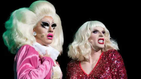 Getty Images Trixie Mattel performs alongside fellow drag queen Katya. Trixie wears her signature enormous blonde wig and exaggerated make-up with a pink jacket over a white blouse. Katya also wears a blonde wig, paired with red lipstick and a sparkly red dress 