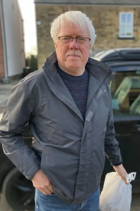 Steve Jones/BBC Paul Gilbert, who has white mid-length hair and a moustache. He is also wearing glasses and holding a white carrier bag.