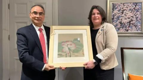 Hu Muqing, wearing a dark suit and red tie, and Laura Pye, wearing a black dress and cream jacket, hold a painting of a white and pink flower in a gold coloured frame during their meeting in China