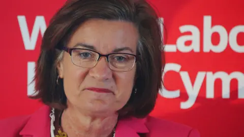 Eluned Morgan speaking in front of a red Welsh Labour banner, wearing a pink jacket. She has a gold chunky necklace on, purple framed glasses and dark brown shoulder-length hair. 