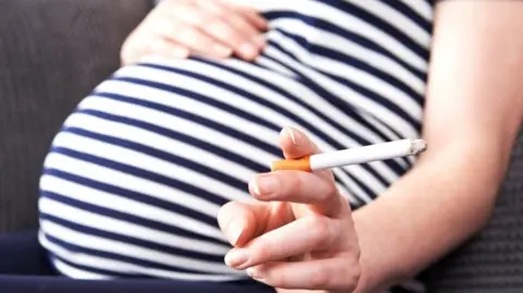 Pregnant woman in blue and white striped top with right hand resting on baby bump and left hand holding a lit cigarette.