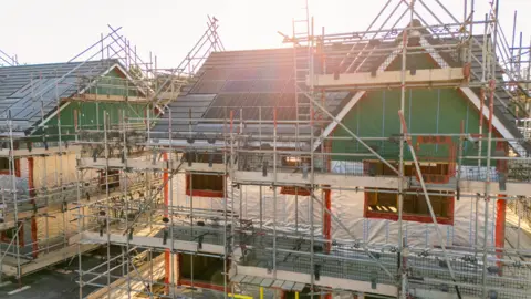 Getty Images Two half-built houses covered in scaffolding. The grey poles of the scaffolding surround the homes which have completed roofs with solar panels on but the bare insulation shows in the walls.