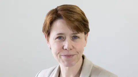 Carers Trust A woman looks at the camera with cropped reddish brown hair, blue eyes and a close-lipped smile. She wears a cream jacket, the headshot is against a white background.