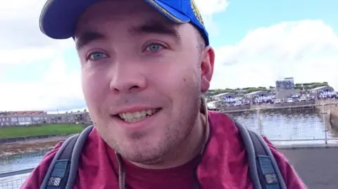 Gerard Coulter is smiling as he stands next to a body of water in front of a stone tower and houses. He wears a blue cap, a scarlet T-shirt and fleece and a rucksack.