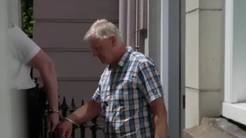 BBC A man being escorted into a van handcuffed to a police officer walking out the back door of a court building.