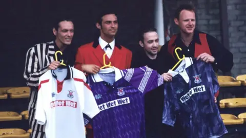 SNS Four men dressed in suits (including one black and white, and one bright red) holding football strips up, coloured white, purple and a dark wavy blue pattern