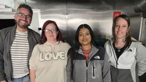 A row of four people looking at the camera - on the left is a man with curly grey hair glasses, a stripy top who is smiling with his teeth. Next is a woman with red hair and glasses wearing a beige jumper with the word 'LOVE.' Next is a woman with dark hair and a North Face jacket on, and on the right is a woman with neck tattoos wearing hoop earrings and smiling with her hands behind her back.