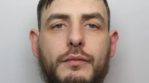 Mugshot of Ashley Pointon in a grey t shirt. He has brown hair and a brown beard.