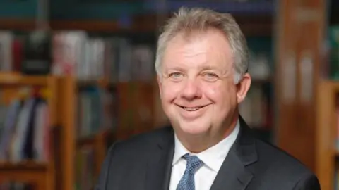 David Sidwick David Sidwick smiles at the camera in a room with bookshelves. He has light grey hair, a dark suit, white shirt and blue tie.