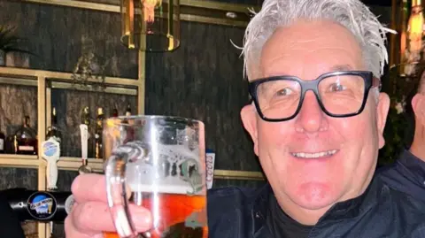 David Thomas with grey hair and wearing a navy blue jacket and black rimmed glasses holds a glass of beer up in a pub. He is smiling.
