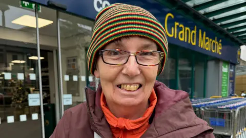 Woman in a stripey hat and glasses standing outside a shopping centre.