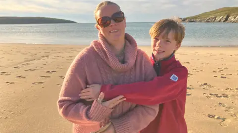 A young boy with blond hair and a red fleece stands of a beach in the sunshine, hugging his mother 