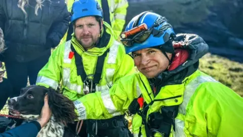 The image shows two people in high-vis jackets, wearing blue helmets and crouching outdoors. There is a black dog with a white speckles in front of them on the left. 