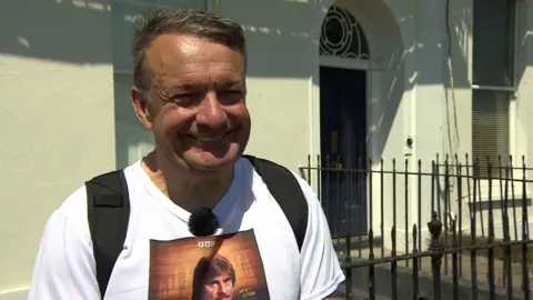 A man with short brown hair wearing a white tee-shirt with a picture of actor Trevor Eve's face on it and a BBC logo. He is standing in front of the railings of a regency property with a fanlight above the door.