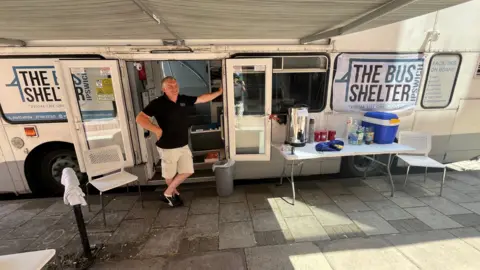 Qays Najm/BBC A bus with its doors open and a man standing by the door. Near the white bus there is a table with beverages including tea and cold drinks.
