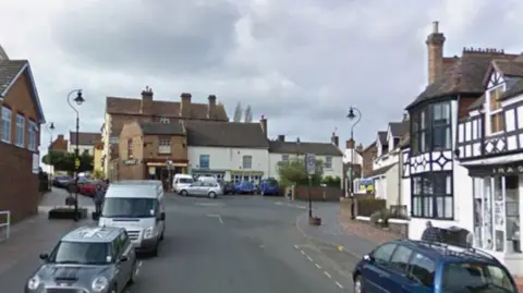 Google A busy street with cars parked on either side and a black and white timber-framed building on the right hand side