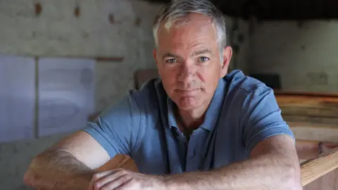 Adam Waugh in the shed where he built his boat, looking into the camera. He has short white hair and blue eyes. He is wearing a blue polo shirt.