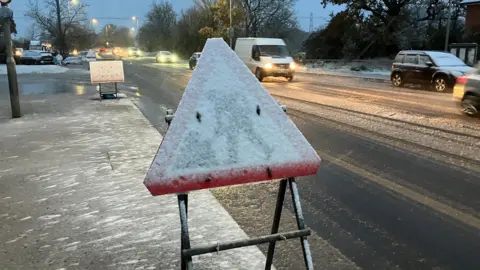 A snow-covered road sign in front of a partially snow-covered road with traffic.