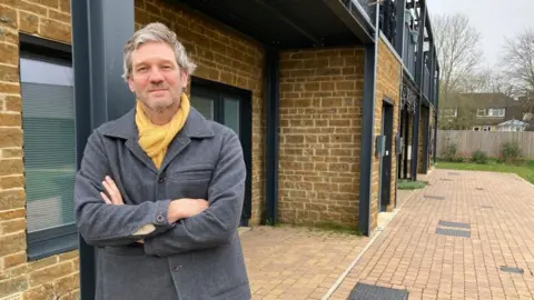 A man is standing, leaning on a pillar. He is wearing a yellow scarf and smiling. Behind him is a brick building. 