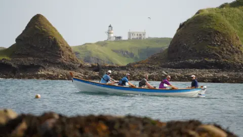 Four rowers and a cox in a boat on the water at Catterline Bay in Aberdeenshire with two hills and lighthouse behind