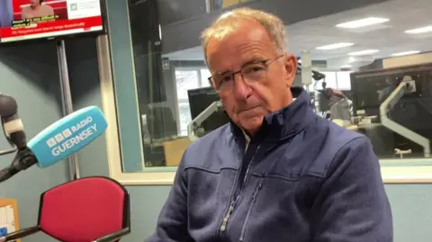 A man sat in a BBC Radio Guernsey studio with a blue microphone in front of him. He is looking straight at the camera. 