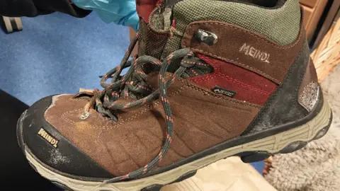 A brown suede walking boot with black and red laces that are undone being held up by a hand wearing a blue rubber glove. 