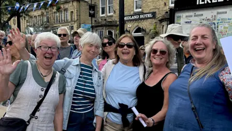Five women smile and wave at the camera