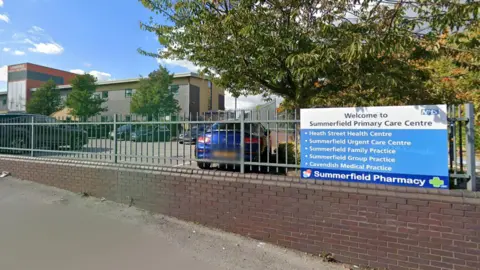 The front of Summerfield Primary Care Centre. There is a blue NHS sign with bullet points on and a sign for the pharmacy. It is on a grey fence and above a brick wall. Behind is a car park and a building. 