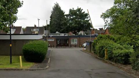 Exterior view of Wednesfield Mortuary, Alfred Squire Road, Wednesfield, Wolverhampton. It has brown front doors and surrounded by bushes and trees. there is a black and yellow striped barrier, which is up.
