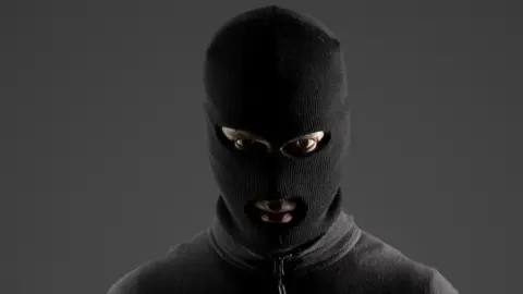 Getty Images A person stares at the camera while wearing a full-face balaclava and a zipped up, grey jumper. They are standing in front of a dark grey wall.