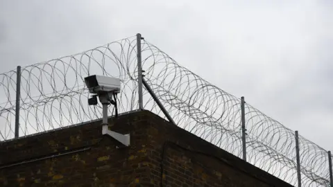 Stock shot of a CCTV camera outside a generic prison. Barbed wire lines the top of a bricked wall.